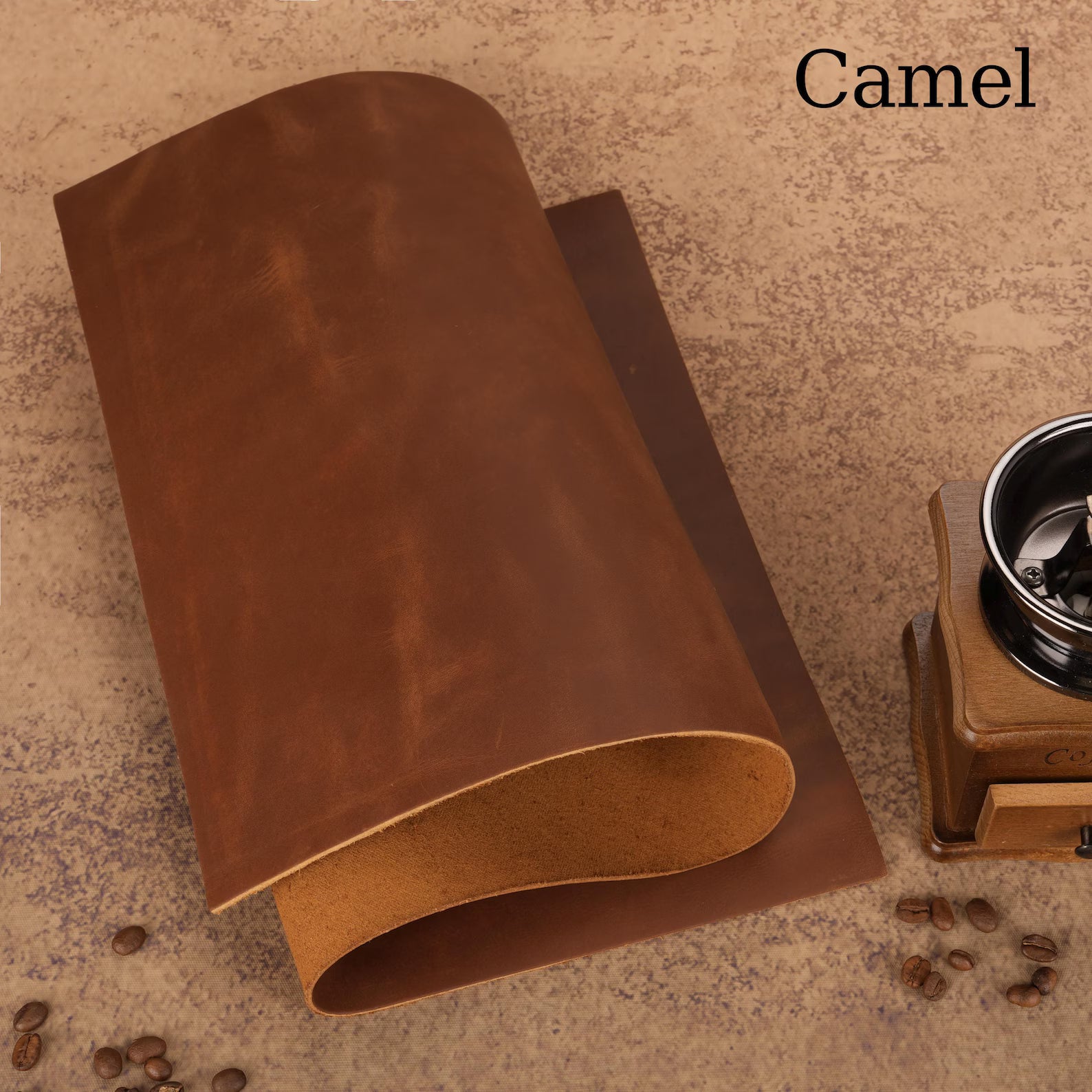 Two pieces of camel-colored leather on a textured surface with coffee beans and a coffee grinder.